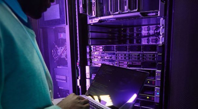 Ingénieur de données afro-américain tenant un ordinateur portable tout en travaillant avec un superordinateur dans la salle des serveurs